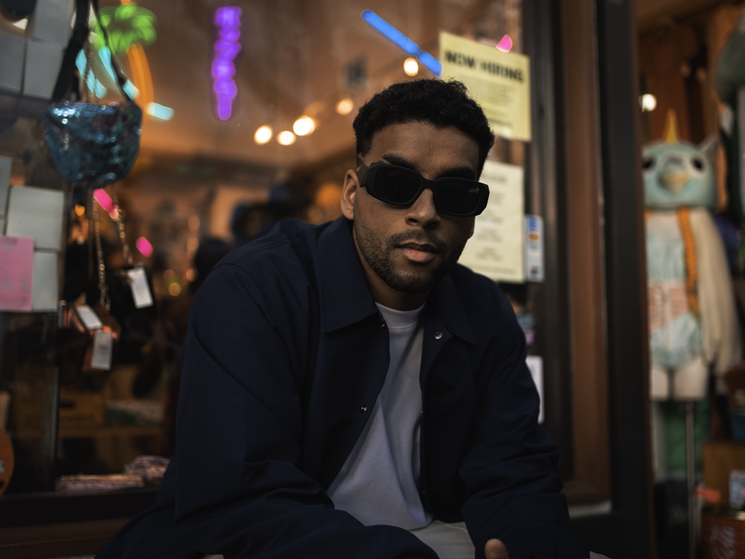 APXO seated in front of a neon-lit storefront at night wearing sunglasses and a dark jacket