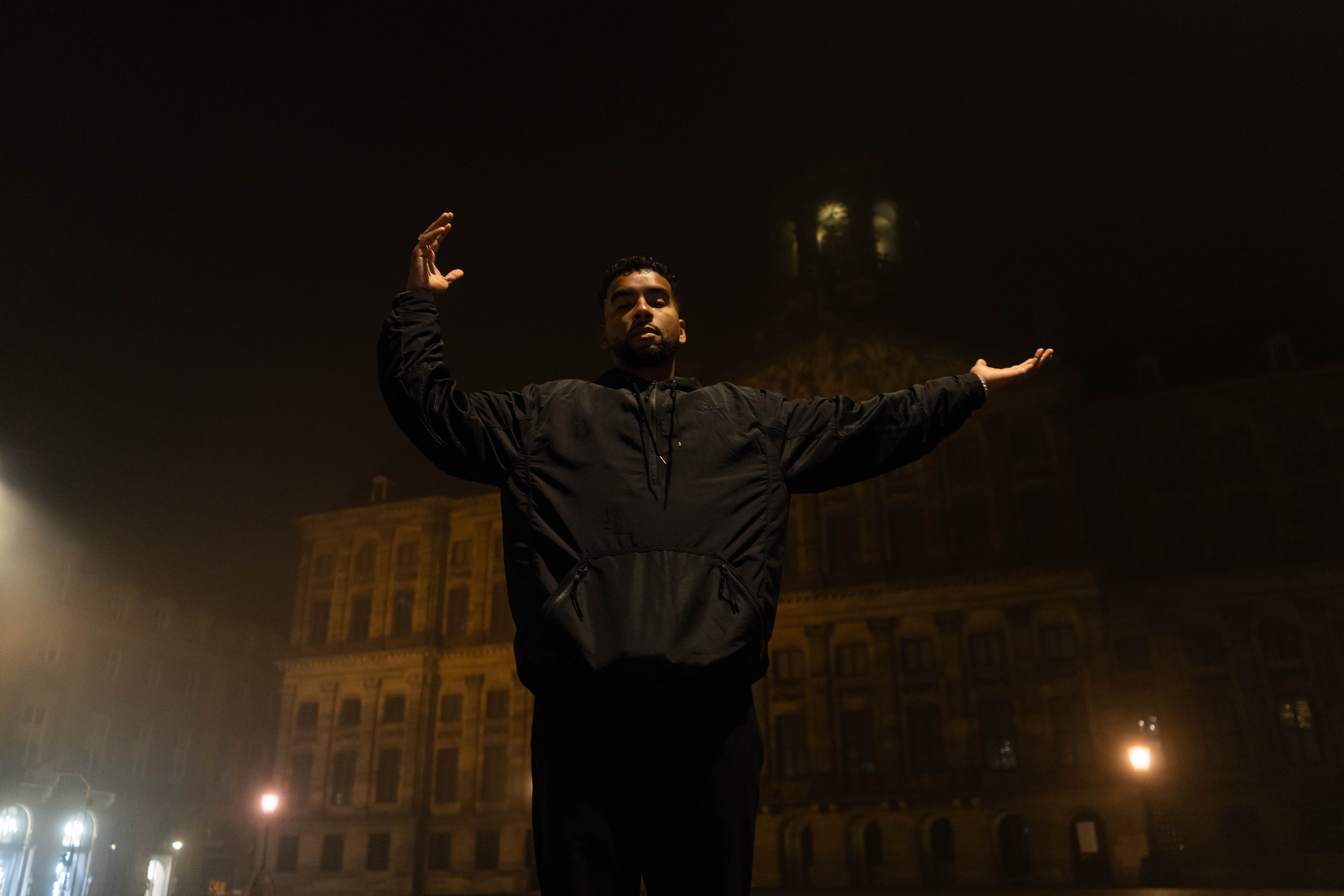 APXO standing with both arms open in a fog-lit European square at night